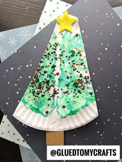 A PAINT SPLAT CRAFTS Christmas tree made from a paper plate, decorated with green paint, red and gold sequins, and topped with a yellow felt star. The tree sits on layered winter-themed papers. @GLUEDTOMYCRAFTS text appears at the bottom.