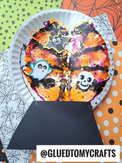 A paper plate painted with vibrant Halloween colors using PAINT SPLAT CRAFTS and decorated with stickers of a bat, ghost, and skull is attached to a black paper base to resemble a bubbling cauldron. Orange, black, and white patterned papers are in the background.