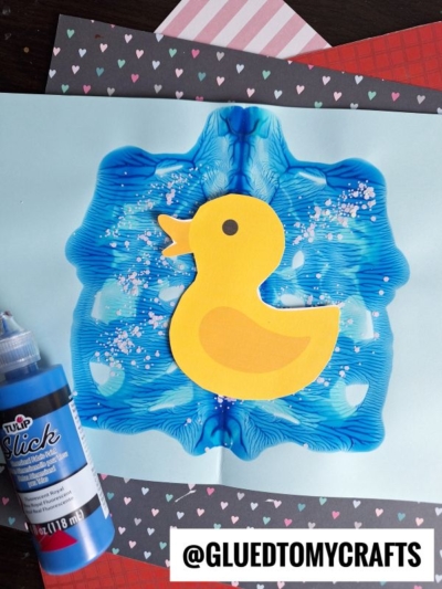 A yellow paper duck is glued on top of blue PAINT SPLAT CRAFTS, smeared in a symmetrical pattern with pink glitter sprinkled on it. Craft supplies and patterned papers surround the artwork.