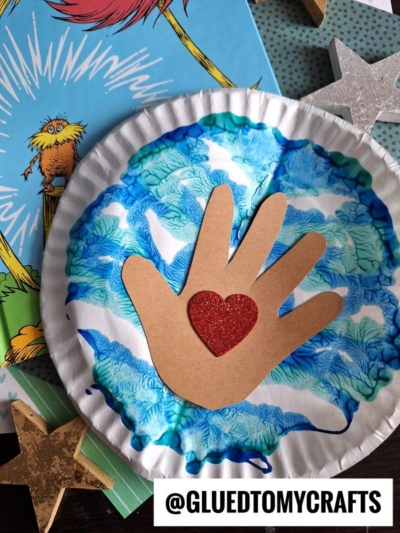 A creative PAINT SPLAT CRAFTS project featuring a paper plate with blue and white patterns, a brown paper hand cutout with a red glitter heart in the center, surrounded by star decorations and a Dr. Seuss book.