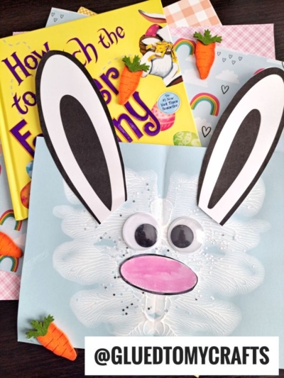 A handmade Easter bunny card with googly eyes, tall ears, and a pink nose is displayed atop children's books, paper carrots, patterned paper, and PAINT SPLAT CRAFTS. @GLUETOMYCRAFTS is written at the bottom.