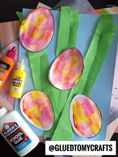 A Spring PAINT SPLAT CRAFTS project features four colorful, oval-shaped paper “flowers” with yellow and pink patterns, green crepe paper stems, a blue paper background, and bottles of glue and paint. The handle @GLUEDTOMYCRAFTS appears at the bottom.