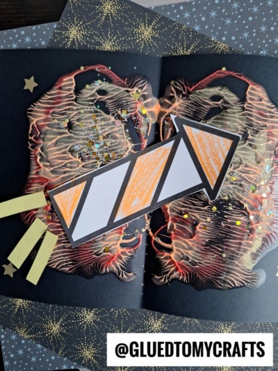 A paper craft showing a rocket over a vibrant, abstract background created with PAINT SPLAT CRAFTS, resembling a nebula or explosion. Star confetti decorates the black paper; the rocket is white with orange sections and yellow strips for flames.
