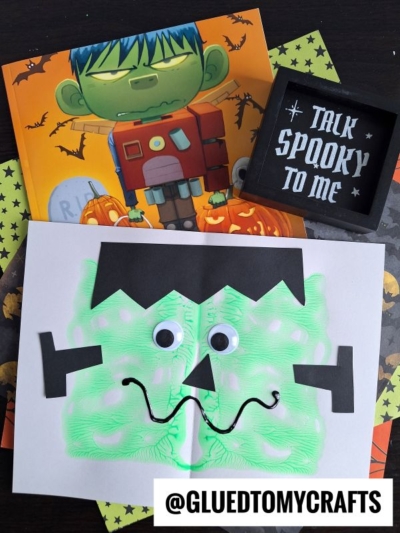 A Frankenstein paint splat craft with googly eyes and black cutouts sits atop a Halloween book and spooky-themed decor. A sign reads "Talk Spooky To Me." @GLUEDTOMYCRAFTS is labeled at the bottom.