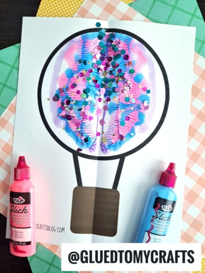 A paper hot air balloon PAINT SPLAT CRAFT with vibrant splatter art inside the balloon and confetti on top. Pink and blue paint bottles sit beside the paper, which rests on a checkered background.