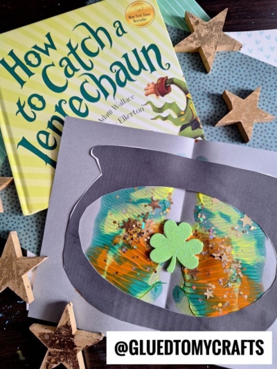 A children’s book titled How to Catch a Leprechaun lies next to a colorful leprechaun-themed PAINT SPLAT CRAFT with a green shamrock, gold glitter, and painted swirls. Wooden stars and confetti are scattered around.