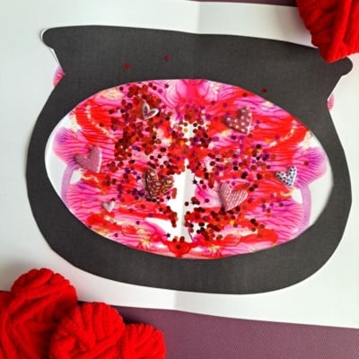 A handmade Valentines Day card features a black fishbowl shape filled with pink, red, and white PAINT SPLAT CRAFTS, glitter, and heart confetti. Red yarn hearts and colorful paper decorate the card. The handle @GLUEDTOMYCRAFTS is visible.