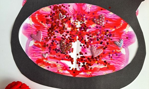 A handmade Valentines Day card features a black fishbowl shape filled with pink, red, and white PAINT SPLAT CRAFTS, glitter, and heart confetti. Red yarn hearts and colorful paper decorate the card. The handle @GLUEDTOMYCRAFTS is visible.