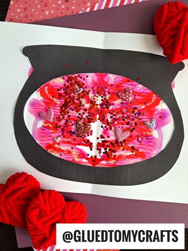 A handmade Valentines Day card features a black fishbowl shape filled with pink, red, and white PAINT SPLAT CRAFTS, glitter, and heart confetti. Red yarn hearts and colorful paper decorate the card. The handle @GLUEDTOMYCRAFTS is visible.