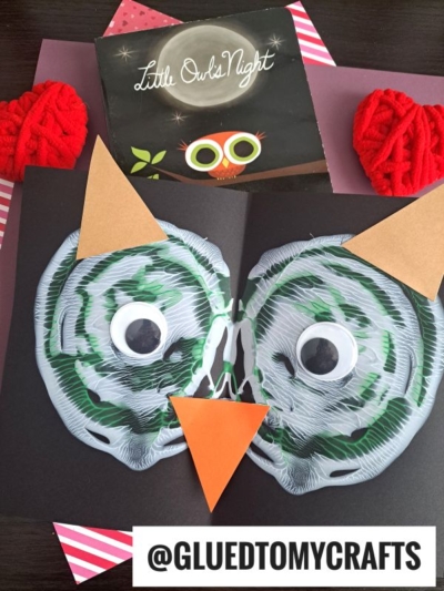 A children's owl PAINT SPLAT CRAFT made from green and white paint on black paper with large googly eyes, plus an orange paper beak and ears. Behind it is the book Little Owls Night and two red yarn balls.
