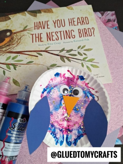 A colorful bird craft inspired by PAINT SPLAT CRAFTS, made from a painted paper plate, blue wings, googly eyes, and an orange beak, sits on craft supplies and the book Have You Heard the Nesting Bird?.