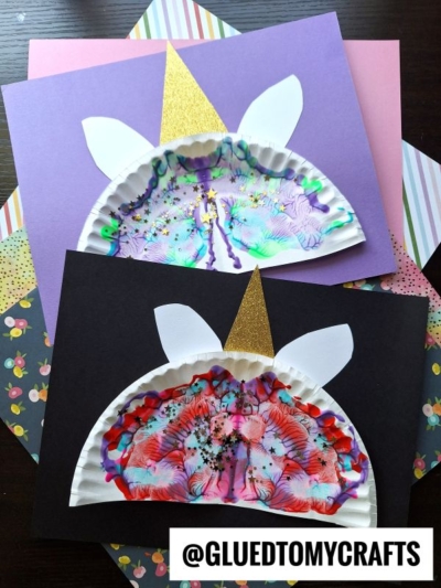 Two unicorn face paint splat crafts made from painted paper plates with glitter, white paper ears, and gold glitter paper horns, displayed on colored paper backgrounds with a @GLUEDTOMYCRAFTS label.