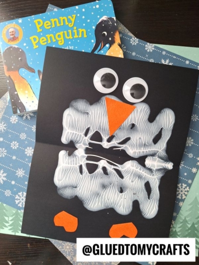 A children’s PAINT SPLAT CRAFT penguin made with black paper, white glue, large googly eyes, and orange paper cutouts for the beak and feet, displayed alongside the book Penny the Penguin on a winter-themed background.