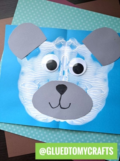 A blue paper features a bear’s face made with a white paint splat, detailed with large googly eyes, gray paper ears, and a paper nose and mouth. Several colorful papers are beneath. Text reads @GLUEDTOMYCRAFTS. Perfect for PAINT SPLAT CRAFTS fun!.