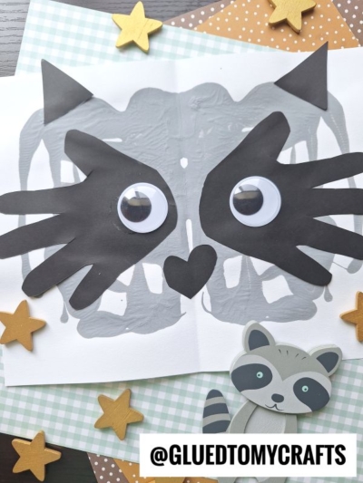 A handmade raccoon face PAINT SPLAT CRAFTS project with gray paint, large googly eyes, and black paper cutouts for ears, mask, and nose, displayed on patterned papers with wooden star decorations and a small raccoon figure.