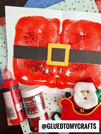 A child’s PAINT SPLAT CRAFTS project shows a red symmetrical painting with glitter, resembling Santa’s suit, decorated with a black belt and yellow buckle. Nearby are red paint, glitter, a Santa ornament, and festive paper.