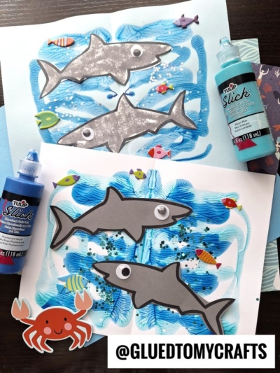 Two colorful shark-themed PAINT SPLAT CRAFTS made from paper and paint are surrounded by bottles of blue craft paint, fish stickers, and a paper crab, displayed on a table with the handle @GLUEDTOMYCRAFTS at the bottom.