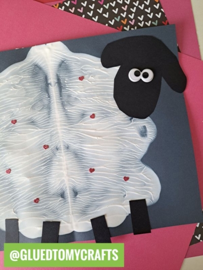 A fun PAINT SPLAT CRAFTS project: a sheep made from white paint spread on dark paper for the body, with black paper legs and a head with googly eyes, placed atop layered pink and heart-patterned papers.