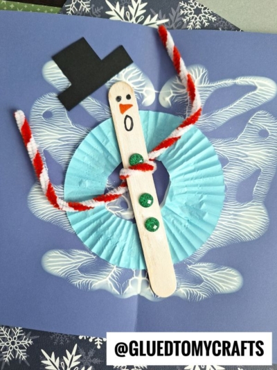 A handmade snowman craft made with a popsicle stick, green buttons, a black hat, a red-and-white pipe cleaner scarf, and a blue cupcake liner, displayed on a blue background with PAINT SPLAT CRAFTS and white designs. @GLUEDTOMYCRAFTS label at the bottom.