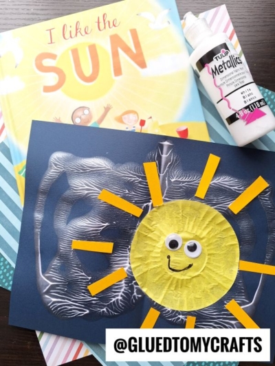 A cheerful children's sun-themed craft project features a yellow sun made from textured paper, yellow rays, googly eyes, and is displayed on dark paper. Behind it are an “I Like the Sun” book and a bottle of white paint for fun paint splat crafts.