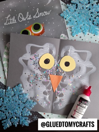A children’s owl PAINT SPLAT CRAFT made with white paint, glitter, yellow paper eyes, and an orange beak sits on a gray background, surrounded by a book, blue glitter snowflakes, and a bottle of glue.