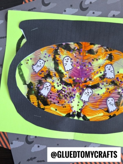 A colorful Halloween PAINT SPLAT CRAFT featuring a black paper cauldron with a swirled orange, yellow, and purple center, decorated with ghost stickers and glitter. The background has Halloween-themed paper. @GLUEDTOMYCRAFTS is labeled at the bottom.