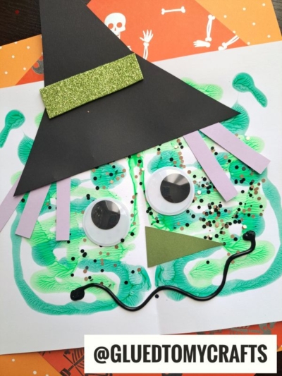 A children’s PAINT SPLAT CRAFT of a witch’s face made with green paint, large googly eyes, sprinkle decorations, a black paper hat with a glittery band, paper hair, and a wavy mouth. The username @GLUEDTOMYCRAFTS is visible.
