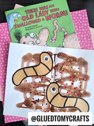A children’s book titled “There Was an Old Lady Who Swallowed a Worm!” sits beside a brown worm craft made of yarn, paper cutouts, and googly eyes, displayed on star-patterned pink papers with playful PAINT SPLAT CRAFTS accents.