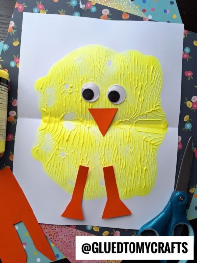 A handmade chick paint splat craft made from yellow paint, two large googly eyes, and orange paper cutouts for the beak and legs, displayed on white paper with colorful patterned paper and scissors nearby.