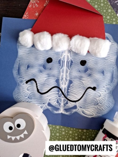 A blue paper features a PAINT SPLAT CRAFTS white symmetrical print resembling a Santa face, with a red paper hat trimmed with cotton balls, drawn eyes and wavy mouth. Craft supplies and papers are scattered around. @GLUEDTOMYCRAFTS is labeled at the bottom.