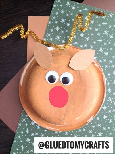 A handmade reindeer craft using a painted paper plate, large googly eyes, a red paper nose, brown paper ears, and a curly gold pipe cleaner antler—perfect for fans of paper bowl crafts—displayed on patterned paper.