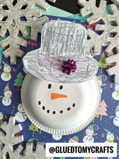 A paper plate snowman craft with a drawn top hat, a purple bow, and an orange carrot nose, surrounded by wooden snowflakes on a festive snowman-patterned background—perfect inspiration for fans of paper bowl crafts. @GLUEDTOMYCRAFTS label at the bottom.