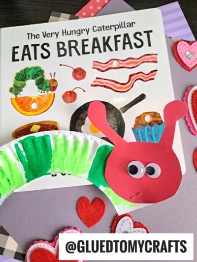 A copy of The Very Hungry Caterpillar Eats Breakfast is surrounded by felt heart decorations and a colorful HUNGRY CATERPILLAR CRAFT made from painted paper, red cardstock, and googly eyes.