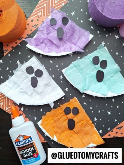 Four ghost crafts made from colored tissue paper and paper plates (purple, teal, white, and orange) with black paper eyes and mouths are displayed on a star-patterned background—fun crepe paper craft ideas with Elmer’s glue and crepe paper rolls nearby.