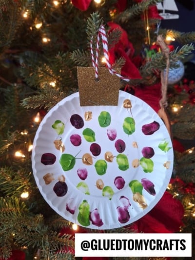A paper plate painted with green, purple, and gold dots hangs on a Christmas tree, decorated to look like an ornament with a glittery gold paper top and red and white string. @GLUEDTOMYCRAFTS is written at the bottom.