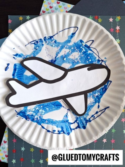 A paper plate decorated with blue paint swirls, inspired by PAINT SPLAT CRAFTS, and a white paper cutout of an airplane, placed on a patterned background. The handle @GLUEDTOMYCRAFTS is displayed at the bottom.