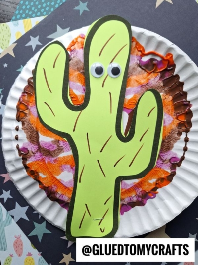 A green cactus cutout with googly eyes is glued onto a paper plate decorated with PAINT SPLAT CRAFTS in orange, brown, and pink. The plate sits on star-patterned paper. @GLUEDTOMYCRAFTS is written at the bottom.