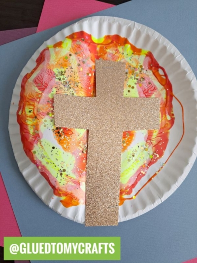 A paper plate decorated with colorful paint swirls in orange, yellow, and pink hues, featuring a large glittery gold cross in the center—a fun idea inspired by PAINT SPLAT CRAFTS. The handle @GLUEDTOMYCRAFTS is displayed at the bottom.