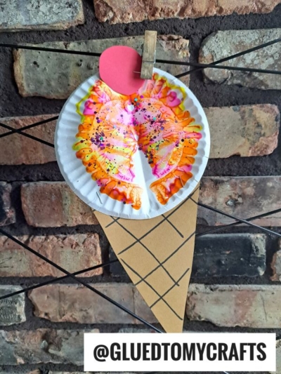 A paper plate painted with colorful, symmetrical paint splat patterns simulates ice cream on a brown paper cone, clipped with a red circle on a string, displayed against a brick wall. Text reads @GLUEDTOMYCRAFTS and PAINT SPLAT CRAFTS.