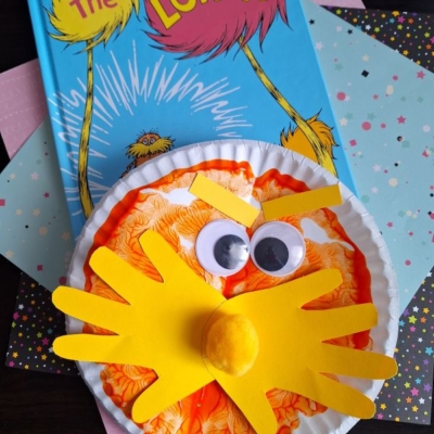 A children’s book, The Lorax, is shown with a paper plate PAINT SPLAT CRAFT of the Lorax featuring orange coloring, googly eyes, a yellow mustache, and nose, displayed on pastel and star-patterned papers.