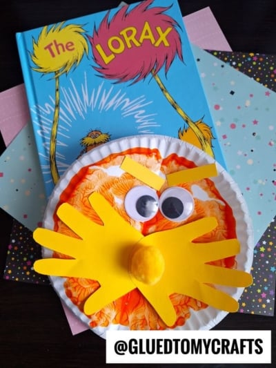 A children’s book, The Lorax, is shown with a paper plate PAINT SPLAT CRAFT of the Lorax featuring orange coloring, googly eyes, a yellow mustache, and nose, displayed on pastel and star-patterned papers.