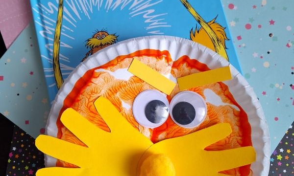 A children’s book, The Lorax, is shown with a paper plate PAINT SPLAT CRAFT of the Lorax featuring orange coloring, googly eyes, a yellow mustache, and nose, displayed on pastel and star-patterned papers.