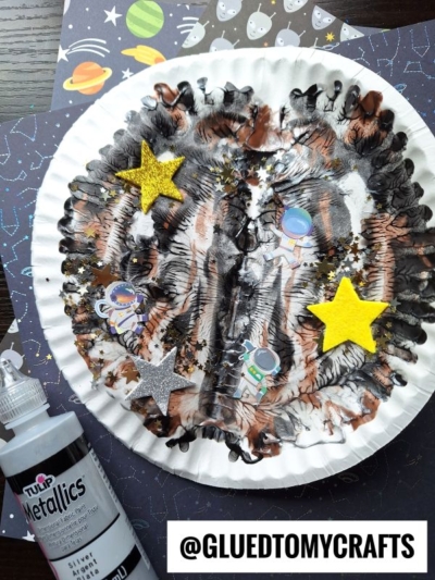 A PAINT SPLAT CRAFTS paper plate decorated with black, white, and copper paint swirls, topped with glitter, astronaut stickers, and star cutouts, sits on a space-themed background next to a bottle of metallic paint.