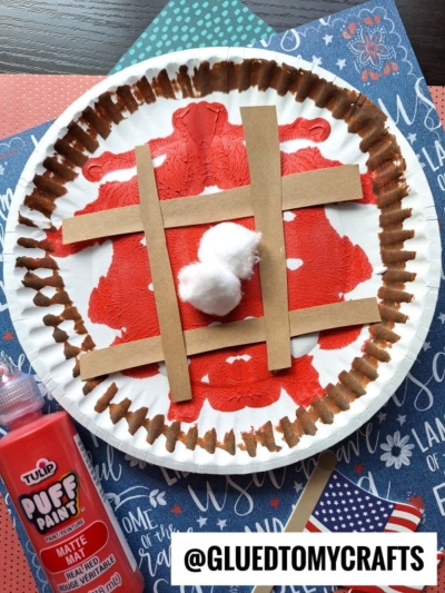A paper plate painted red in the center, like a PAINT SPLAT CRAFT, with brown paper strips as a tic-tac-toe grid and two cotton balls in the middle. Craft supplies and patterned paper decorate the background.