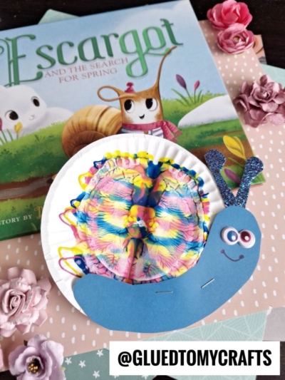 A colorful snail craft made from a white paper plate and blue paper, featuring rainbow-patterned cupcake liners for the shell—perfect for pairing with Escargot and surrounded by pink paper flowers, it’s an inspiring addition to any PAINT SPLAT CRAFTS collection.