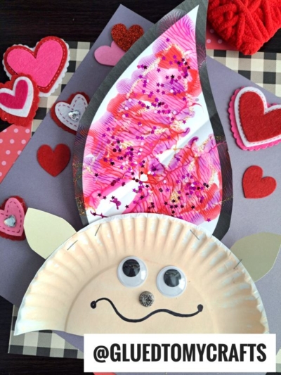 A paper plate PAINT SPLAT CRAFT shaped like a gnome, featuring a pink and red painted hat, googly eyes, a smiling face, heart decorations, and the text @GLUEDTOMYCRAFTS at the bottom.