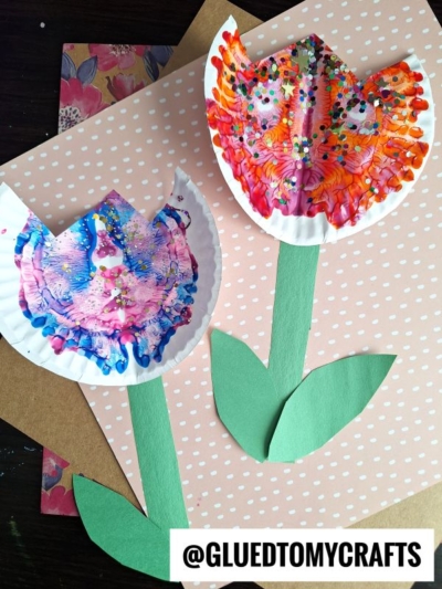 Two tulip flowers made from painted paper plates with vibrant paint splat crafts, attached to green paper stems and leaves, displayed on a pink polka dot background. @GLUEDTOMYCRAFTS is written at the bottom.