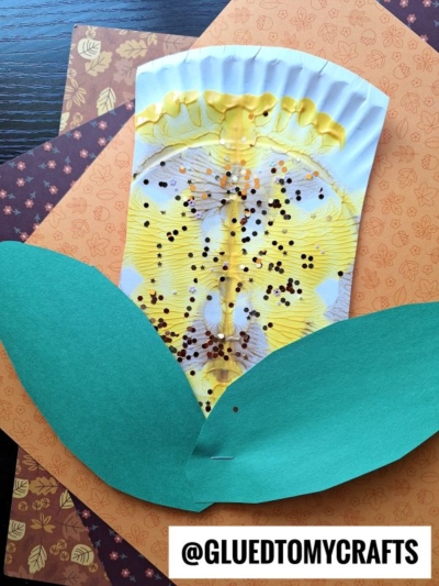 A handmade flower craft inspired by PAINT SPLAT CRAFTS, featuring a yellow painted coffee filter with glitter, green paper leaves, and autumn-themed background papers. The text “@GLUEDTOMYCRAFTS” appears at the bottom.
