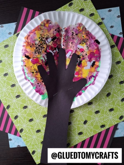 A colorful autumn tree craft inspired by paint splat crafts, made with a paper plate for the leafy top and a black paper hand cutout for the trunk, displayed on patterned papers with spider and stripe designs.