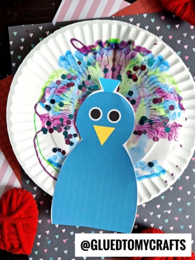 A blue paper peacock with a yellow beak and round eyes is attached to a colorful, paint-splattered paper plate—a perfect example of PAINT SPLAT CRAFTS—set on a patterned background with red pom-poms and heart motifs.